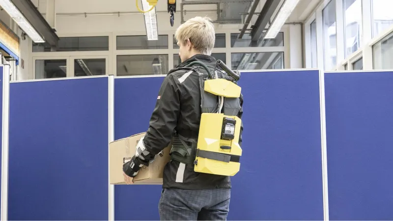 Eine Person ist von vorne zu sehen, die einen Aluträger hebt und dabei ein Exoskelett trägt. Das Exoskelett unterstützt dabei aktiv die Arme beim Heben und Halten des Aluträgers. Dabei ist von vorne nur das Bedienpanel, Rucksacktragegurte und die Seile zu den Armen sichtbar.